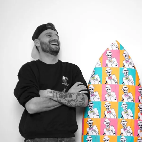 Black and white photo of James next to a surfboard. The surfboard is the only object in the photo that is shown in colour.